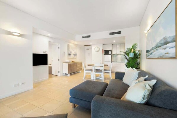 A modern living room with a grey sofa, a flat-screen TV, and a dining area with a white table and chairs.