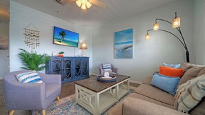 A living room with a sofa, armchair, coffee table, and a cabinet with glass doors. There are wall-mounted paintings and a ceiling fan. - 4 Costa Bahia Condos (Port Aransas)
