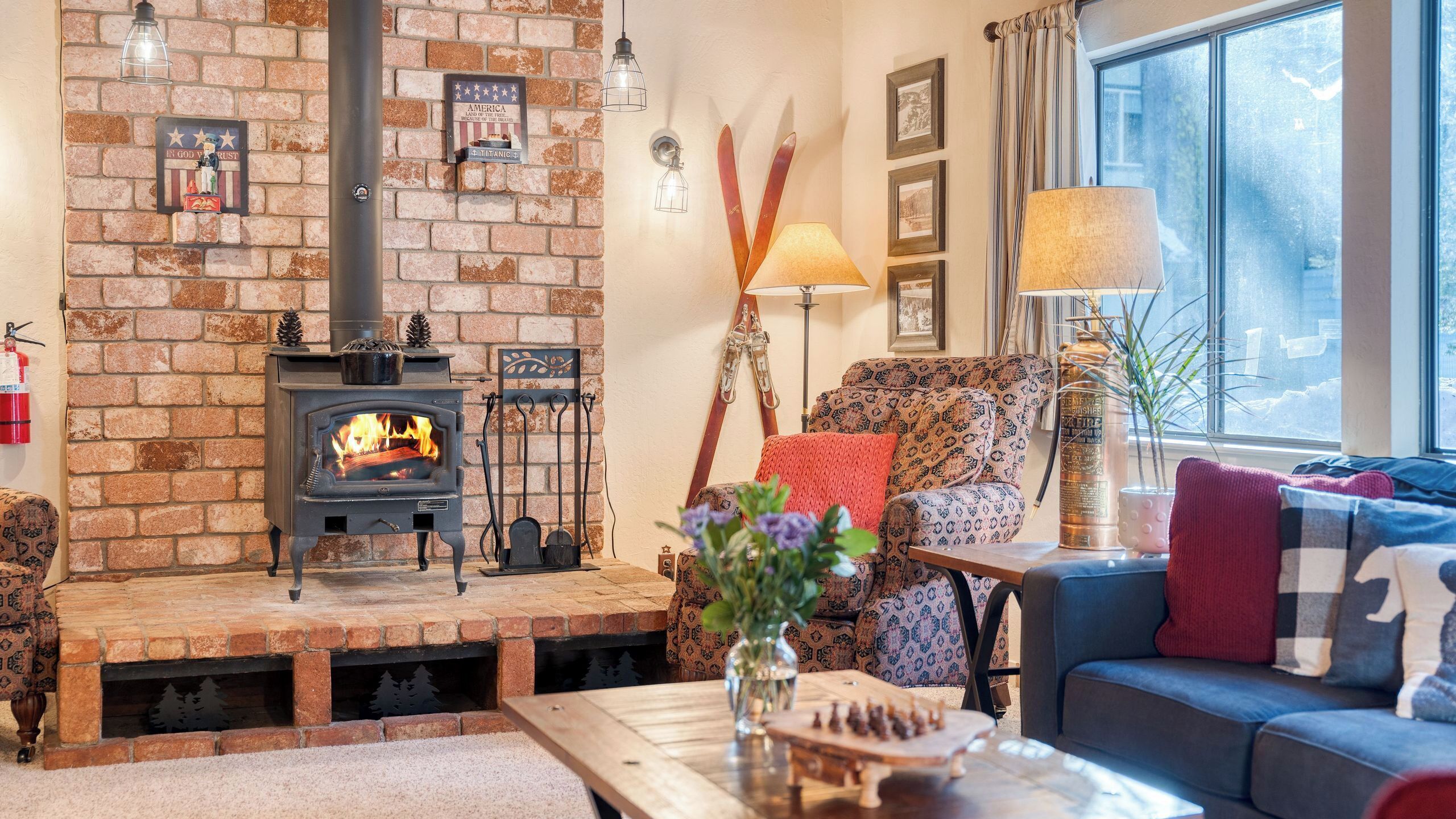 A cozy living room with a brick fireplace, a sofa, a table with a vase, and a window with curtains.