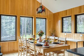 A wooden-paneled dining area with large windows, a table set for breakfast, and a colorful hanging light fixture. - Rustic Arch Cape Getaway w/ Vibrant Interior Walk to the Beach Cape Wonder (Arch Cape)