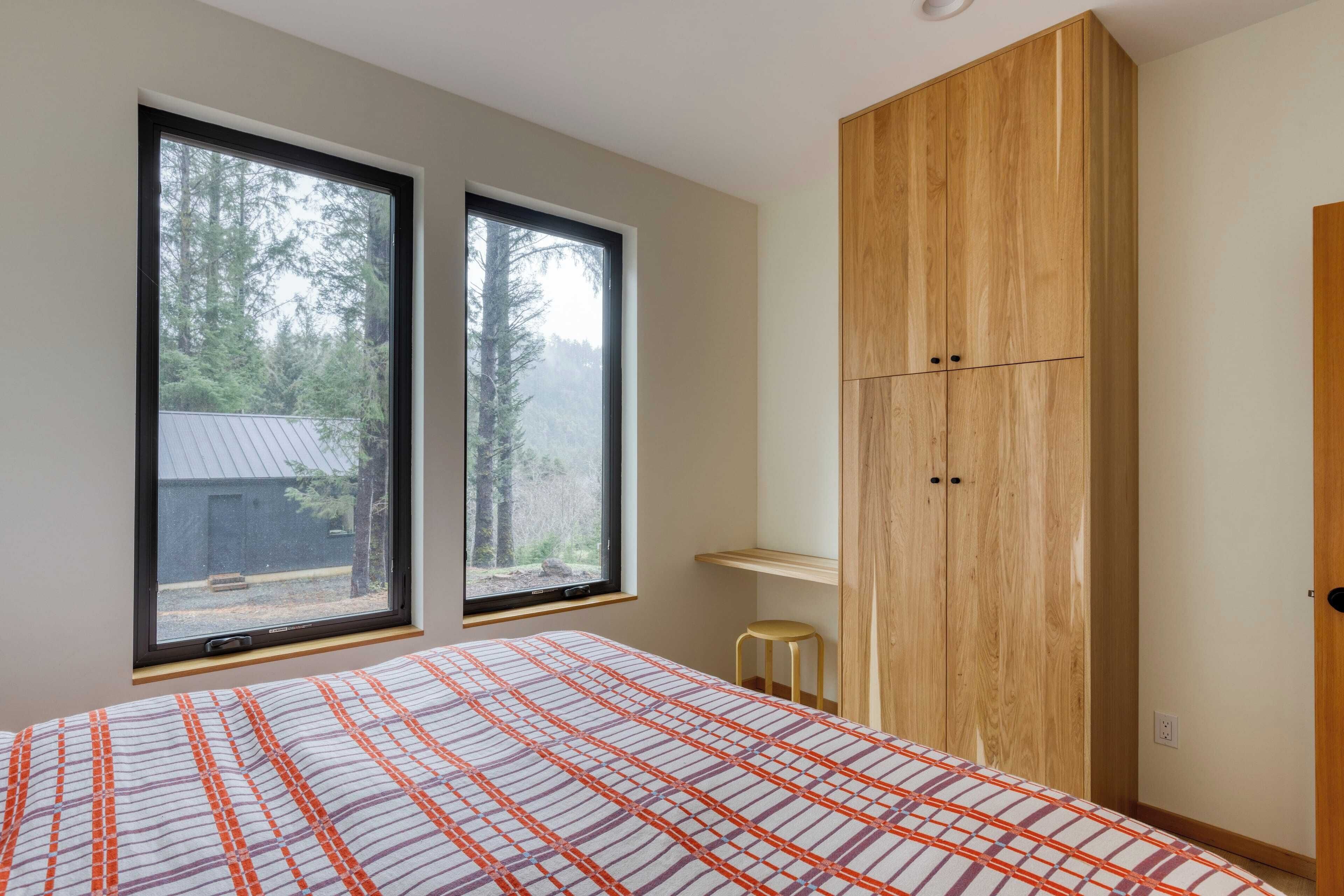 Un dormitorio con cama, dos ventanas, un armario de madera y un taburete.