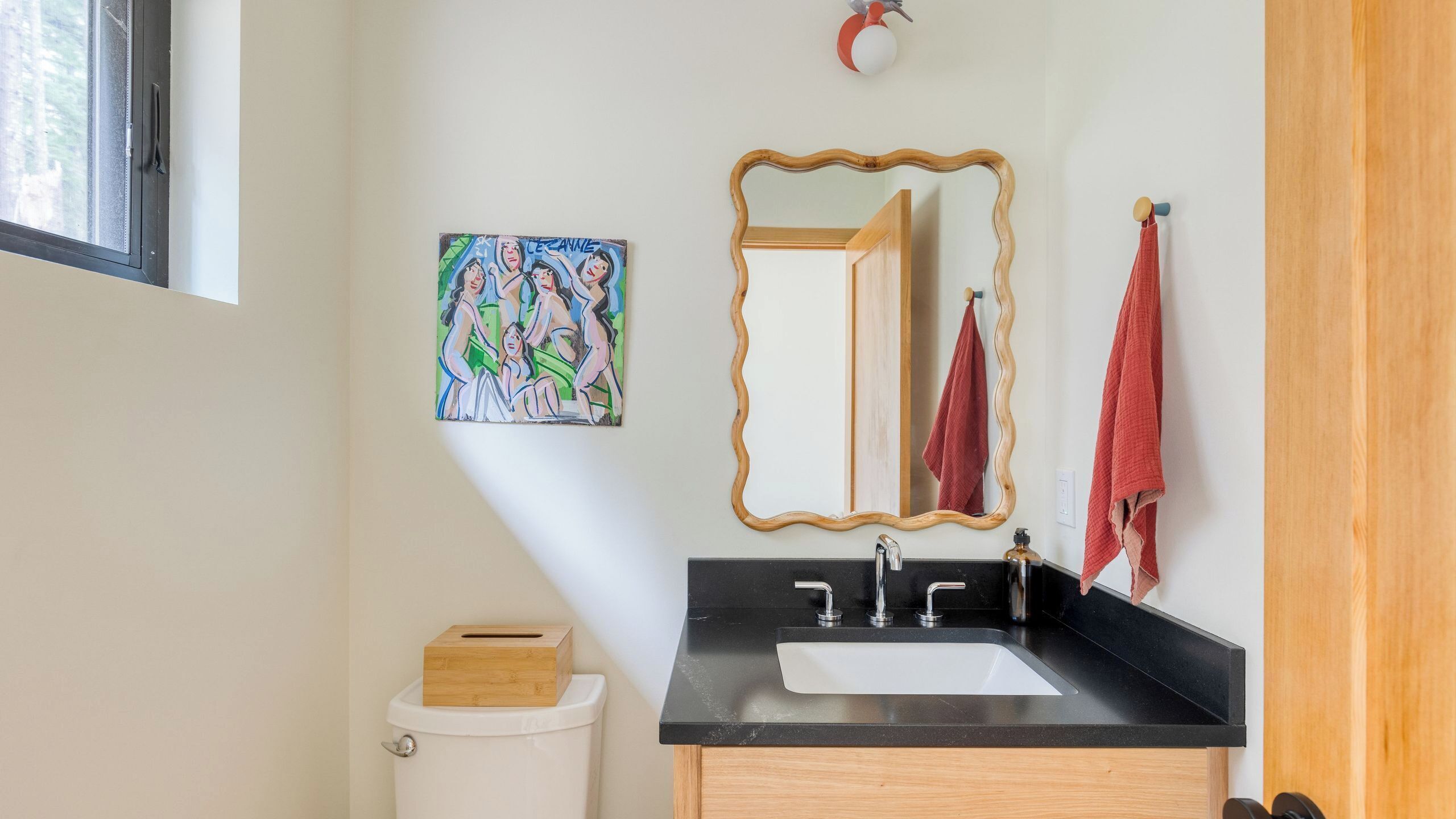 A bathroom with a black sink, wooden vanity, a mirror with a wooden frame, a toilet, and a painting on the wall.