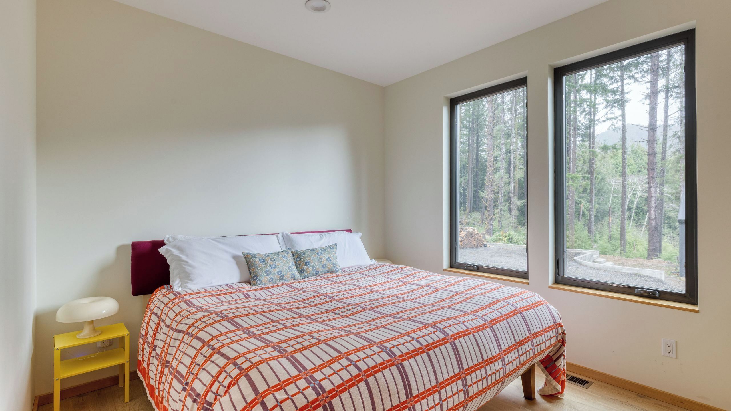 A bedroom with a bed, two windows, a bedside table, and a view of a forest.