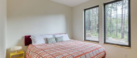 A bedroom with a bed, two windows, a bedside table, and a view of a forest.