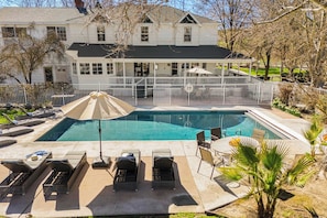 A swimming pool with lounge chairs and umbrellas, surrounded by a white fence and a house in the background. - Victorian Manor In Paso Robles Wine Country + Pool, Hot Tub, Firepit, Tennis Court Sleeps 28 (Paso Robles)