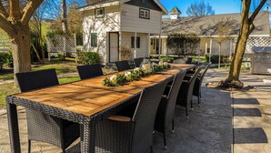 An outdoor dining area with a long wooden table and wicker chairs, surrounded by trees and a house. - Victorian Manor In Paso Robles Wine Country + Pool, Hot Tub, Firepit, Tennis Court Sleeps 28 (Paso Robles)