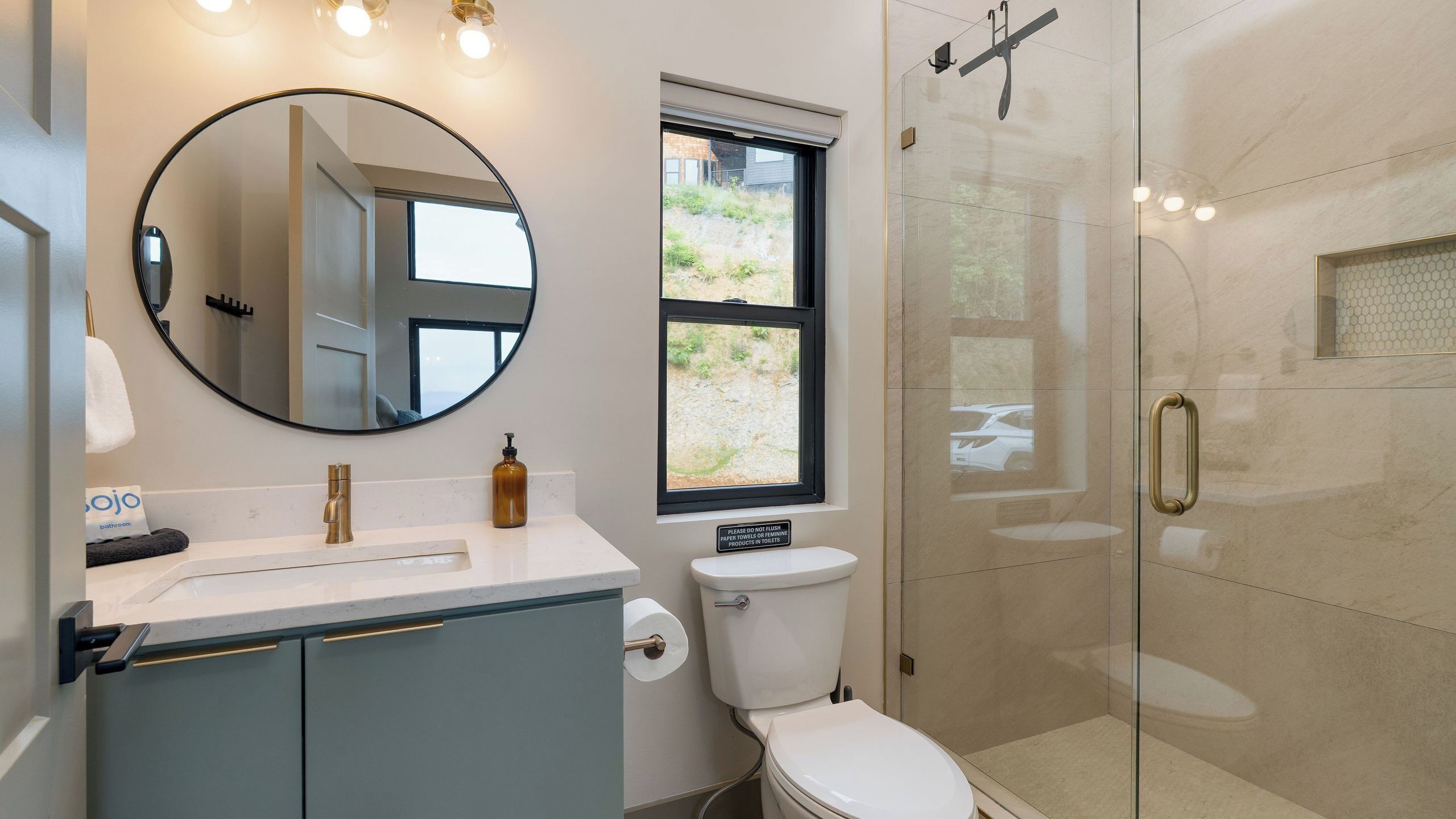 A bathroom with a round mirror, a toilet, a sink, a shower, and a window.