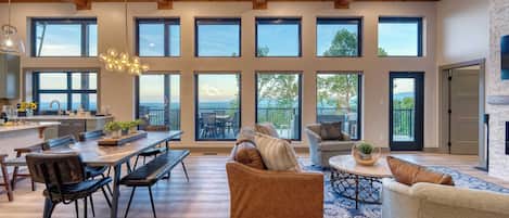 A spacious living area with a large wooden dining table, multiple seating options, and large windows offering views of the outdoors.