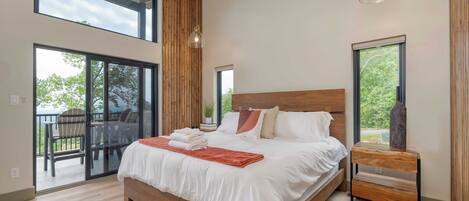 Un dormitorio moderno con una cama grande, cabecera de madera y una mesita de noche con un florero. Hay ventanas amplias y un balcón con mobiliario exterior.