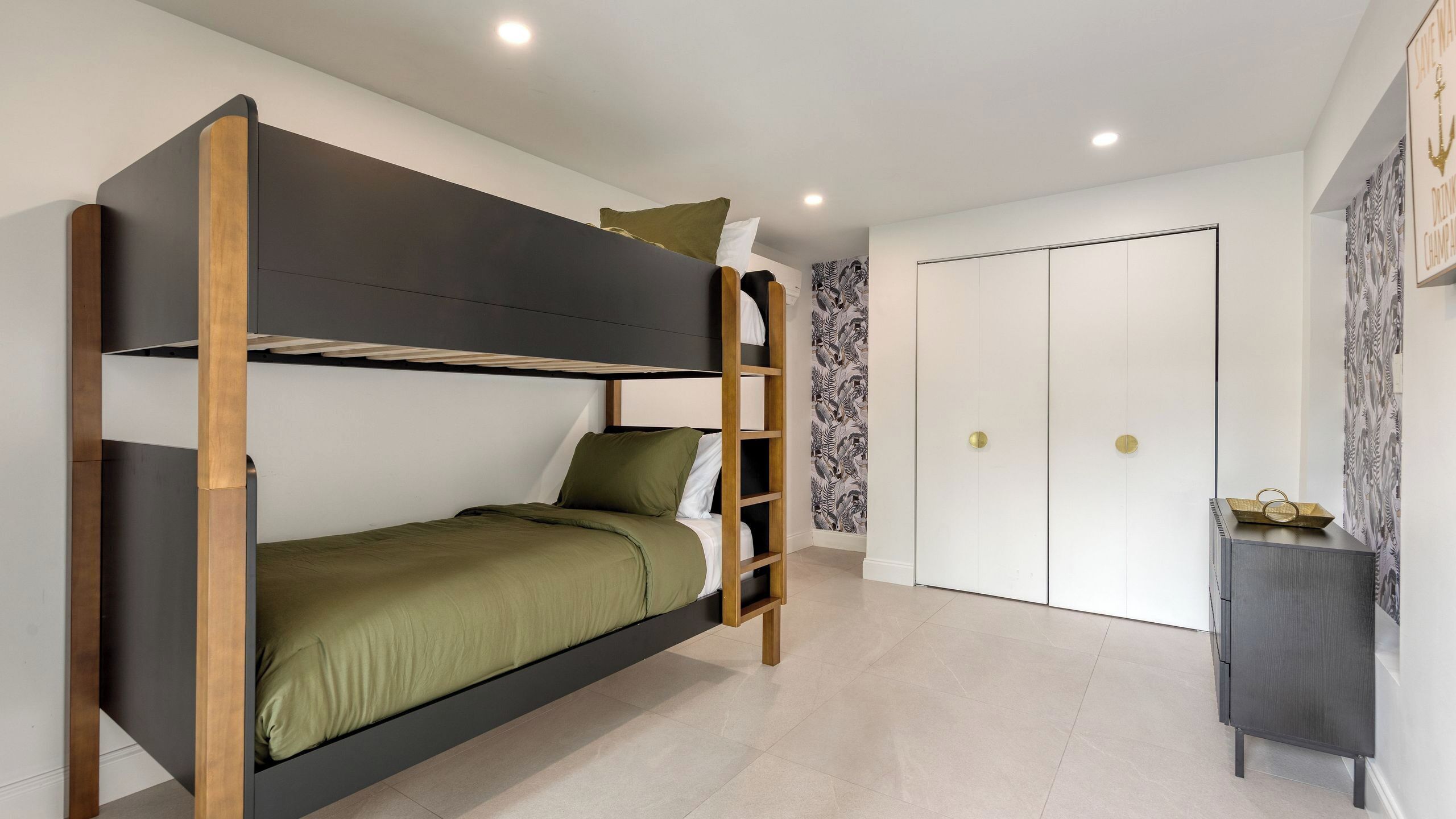 Una cama individual con escalera, un pequeño armario y una puerta en una habitación con papel tapiz estampado y suelo de color claro.