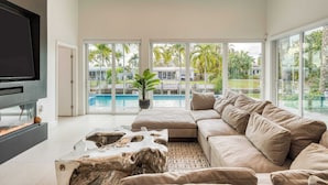 A modern living room with a large sofa, a flat-screen TV, and a view of a swimming pool and palm trees. - Luxury Waterfront Home w/ Pool, Hot Tub & Dock Near Las Olas Gulfstream (Fort Lauderdale)