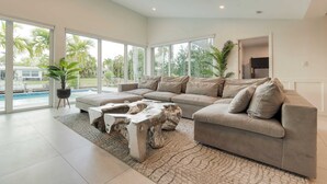 A modern living room with a sectional sofa, a unique coffee table, and large windows offering a view of palm trees and a swimming pool. - Luxury Waterfront Home w/ Pool, Hot Tub & Dock Near Las Olas Gulfstream (Fort Lauderdale)