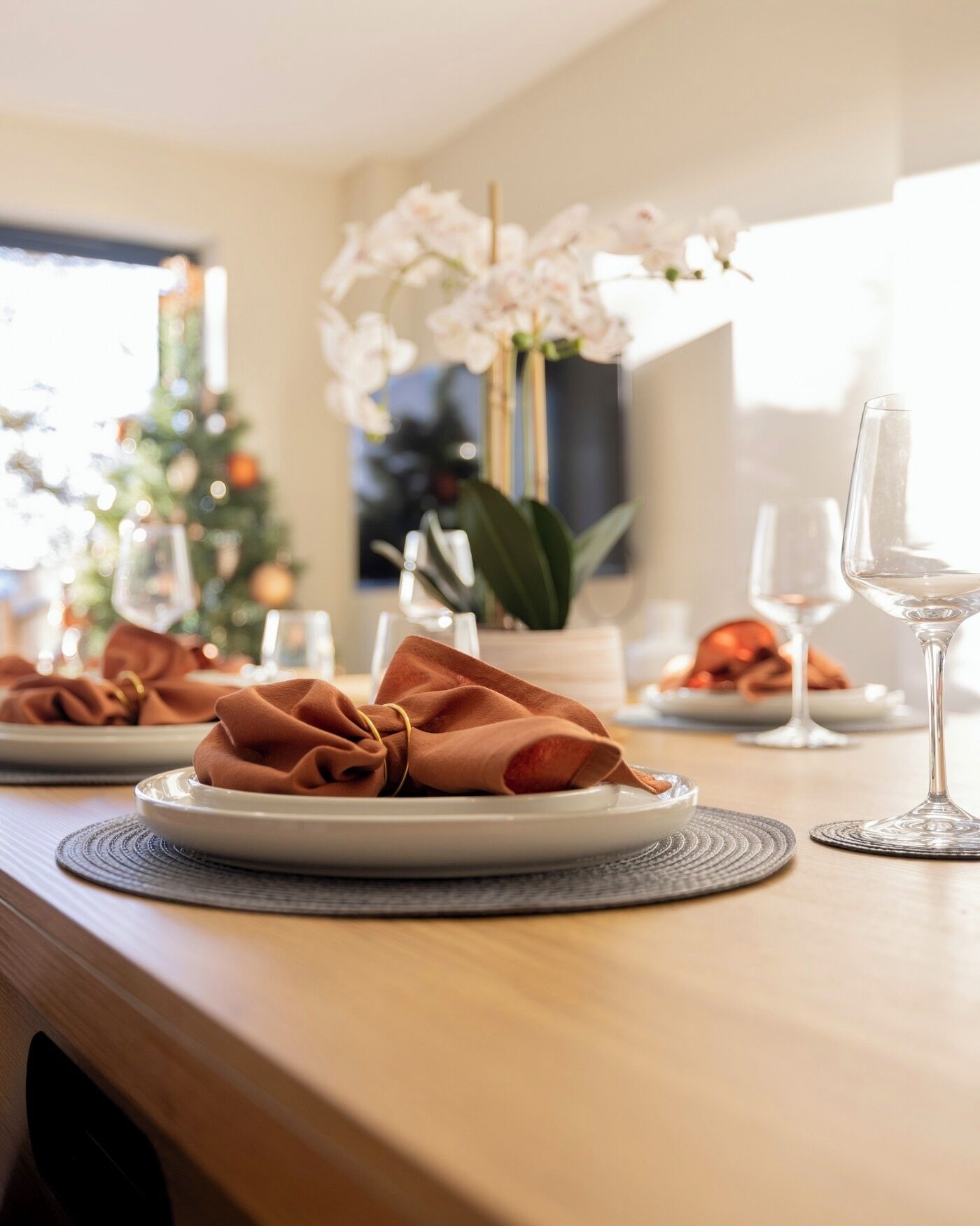 A dining table set with plates, napkins, and wine glasses, with a Christmas tree and flowers in the background.