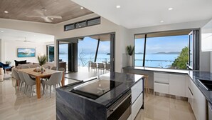 A modern kitchen with a large island, a dining area with a view, and a living space with a sofa and a painting. - Hidden Cove 2 - 4 Bedroom, 4 Bathrooms, Waterfront with Private Pool and buggy (Hamilton Island)