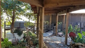 Un espacio al aire libre cubierto con un sendero de piedra, pilares de madera y una terraza de madera con barandilla.