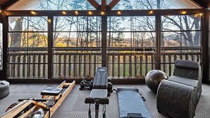 A gym with exercise equipment, a bench, and a chair, overlooking a wooden deck and trees. - Sleeps 13 | Pet-Friendly, Hot Tub, Sauna & Theater (Sevierville)