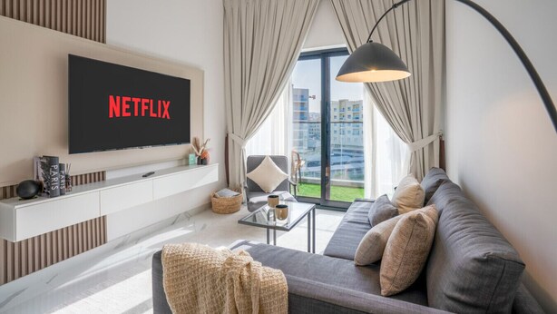 Ein modernes Wohnzimmer mit einem Flachbildfernseher, der 'NETFLIX' anzeigt, einem Sofa, einem Couchtisch und Blick auf ein Gebäude durch große Fenster.