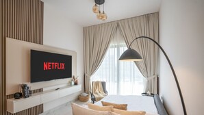 Ein modernes Hotelzimmer mit großem Flachbildfernseher, der 'NETFLIX' anzeigt, einer Couch, einem Nachttisch mit Büchern, einer Stehlampe und einem großen Fenster mit Vorhängen.
