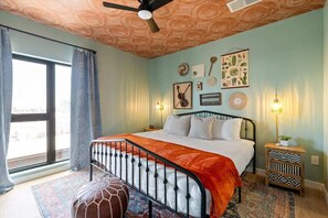 Une chambre à coucher avec un lit, un ventilateur de plafond, une fenêtre avec des rideaux, un mur orné de tableaux encadrés et une table de chevet avec une lampe.
