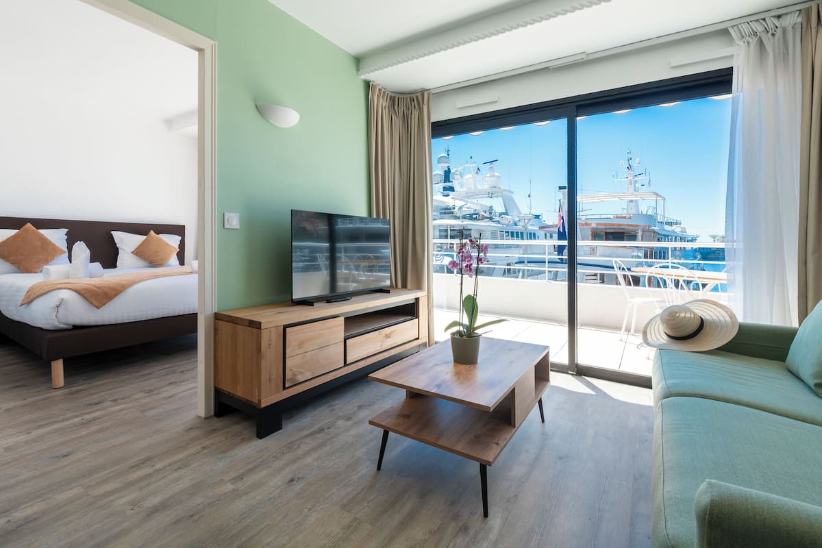 Habitación de hotel moderna con cama, televisor, una mesita de centro de madera, sofá y un ventanal con vistas a un yate.