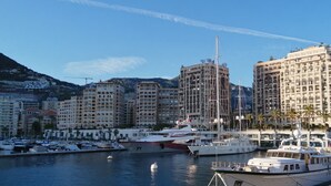 A marina with yachts docked, surrounded by multi-story buildings with balconies and a mountainous backdrop.
