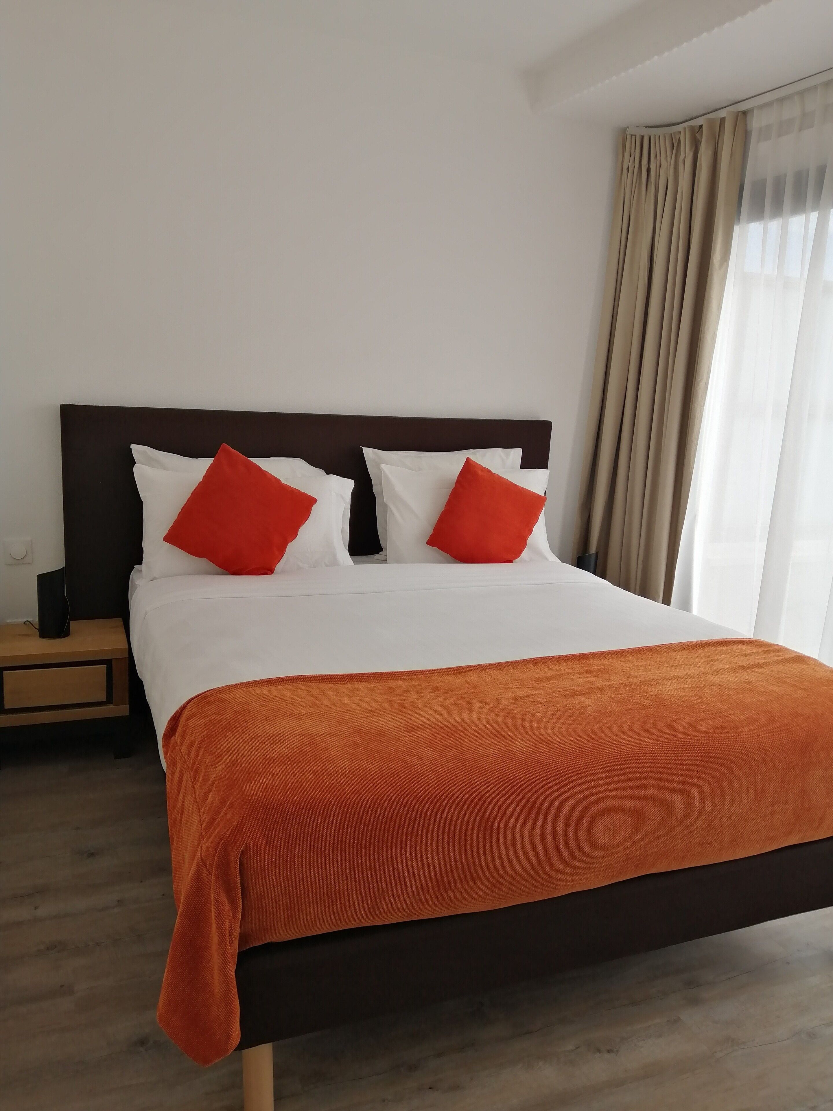 Ein ordentlich bezogenes Bett mit weißer und orangefarbener Bettwäsche, ein Nachttisch aus Holz mit einem Lautsprecher und ein Fenster mit beigen Vorhängen.