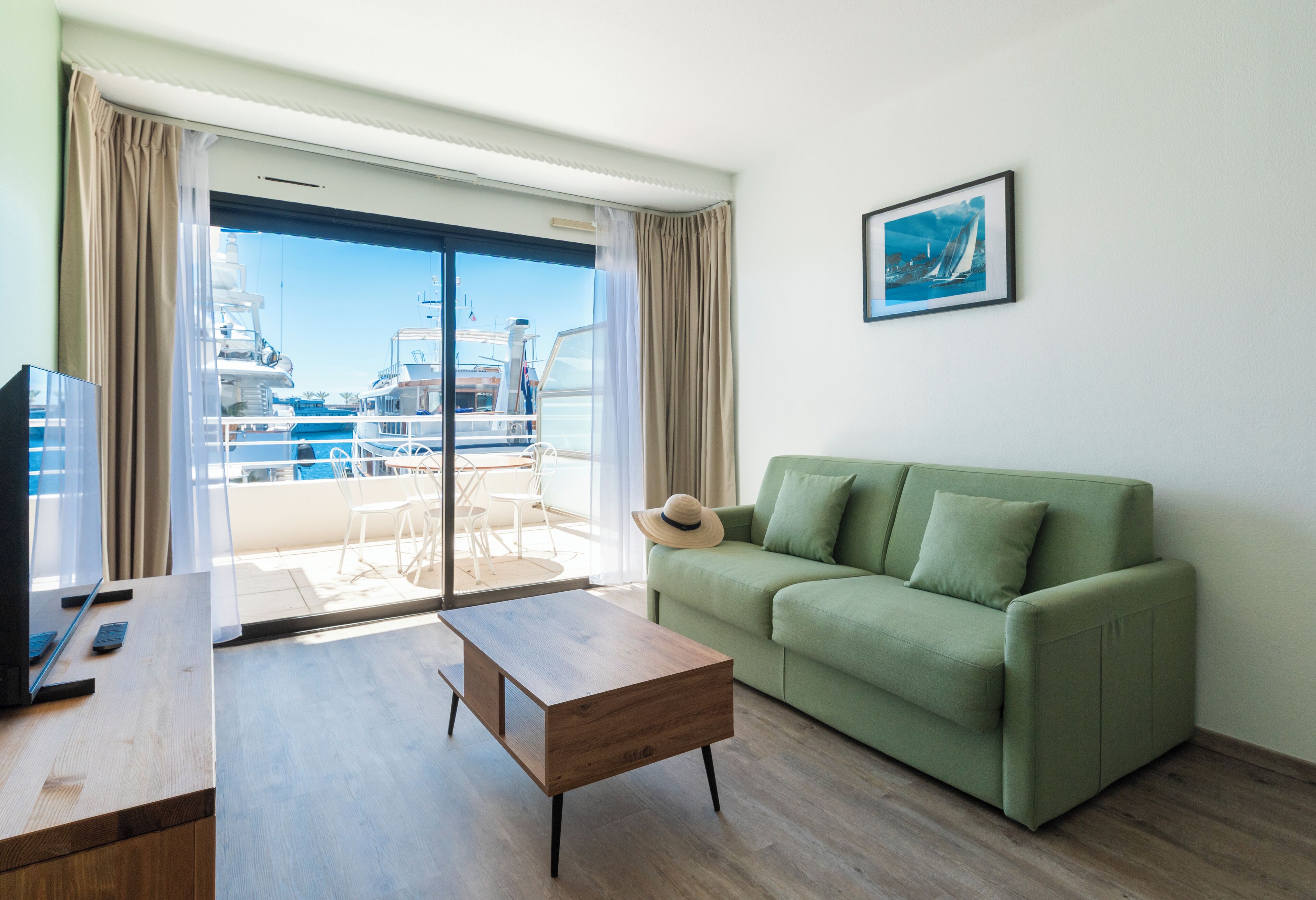 A living room with a green sofa, a wooden coffee table, a TV, and a large window with a view of boats and a marina.