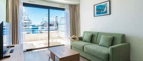 A living room with a green sofa, a wooden coffee table, a TV, and a large window with a view of boats and a marina.