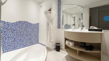 Ett modernt badrum med ett vitt badkar, en vägg täckt med blå mosaikplattor, en stor spegel och ett handfat med hyllor.
