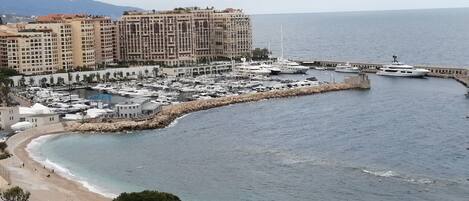 A coastal marina with yachts docked, a beach, and a large hotel building in the background.