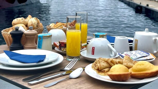 A table with breakfast items including bread, juice, and coffee, set against a backdrop of a boat and canal.