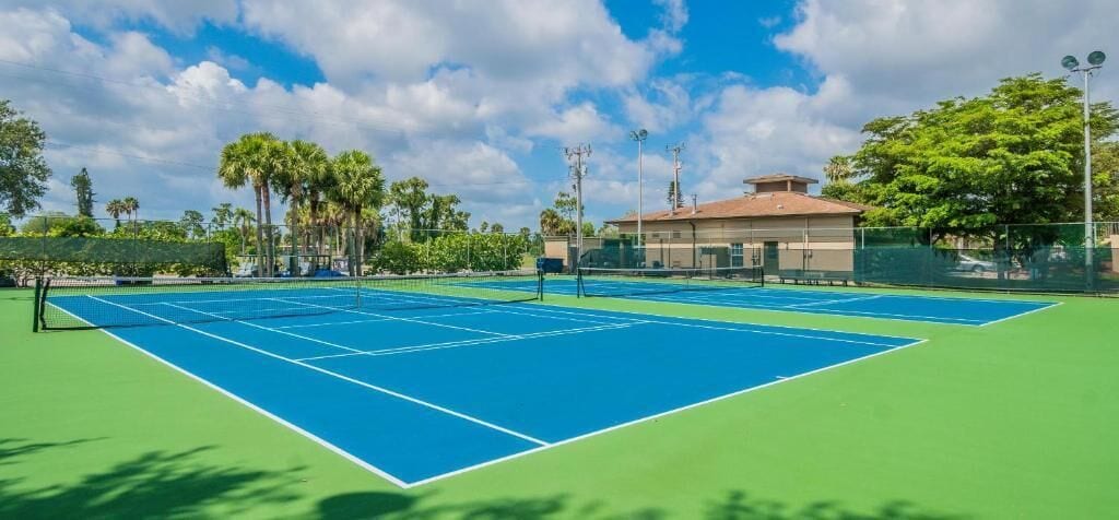 An outdoor tennis court with a net, surrounded by a fence and palm trees.