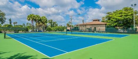 An outdoor tennis court with a net, surrounded by a fence and palm trees.