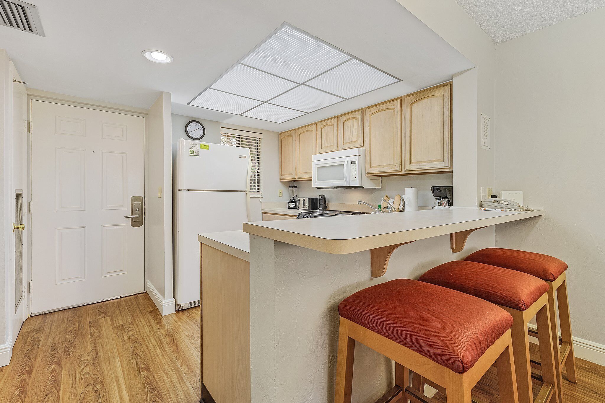Cocina compacta con refrigerador blanco, microondas, lavamanos y muebles de madera. Hay dos taburetes rojos en la barra.