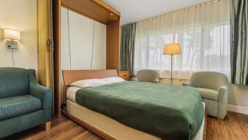 A hotel room with a bed, a green sofa, a patterned armchair, a nightstand, and a lamp.