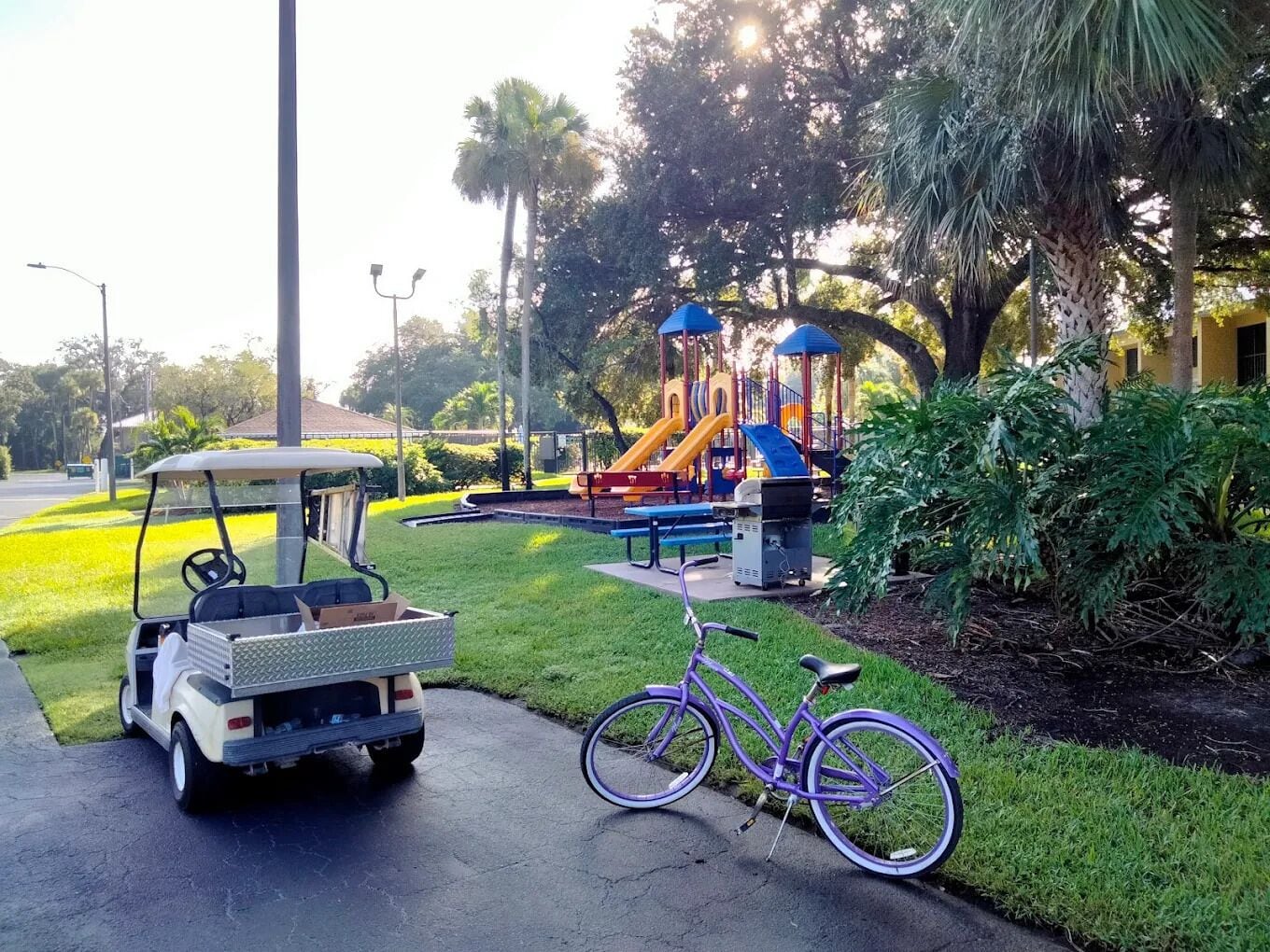 Un carrito de golf estacionado en un camino pavimentado con una bicicleta cerca, un parque infantil con toboganes y estructuras de escalada, y abundante vegetación al fondo.