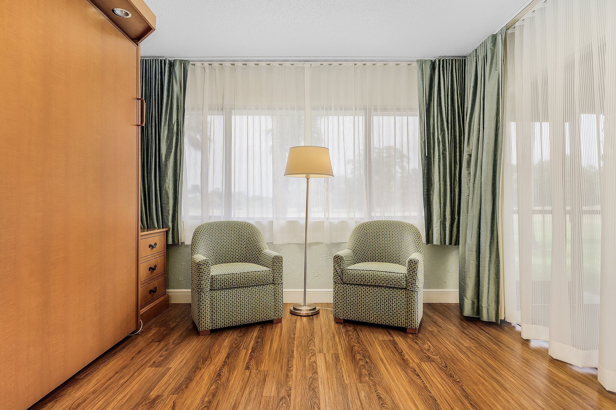 A room with a wooden floor, two patterned armchairs, a floor lamp, and a large window with sheer curtains.