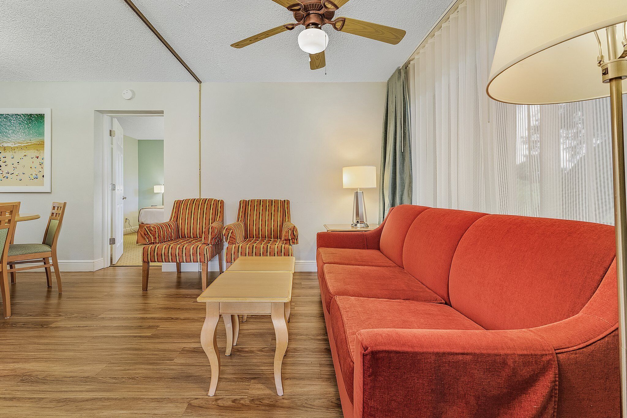 A living room with a wooden floor, a red sofa, striped armchairs, a coffee table, a ceiling fan, and a lamp.