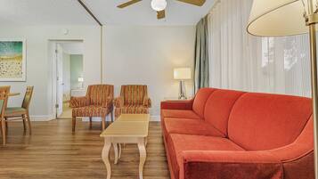 A living room with a wooden floor, a red sofa, striped armchairs, a coffee table, a ceiling fan, and a lamp.