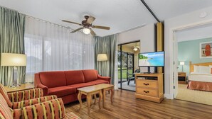 Quarto de hotel com sofá vermelho, poltronas listradas, mesa de centro de madeira, televisão, cômoda de madeira e cama com vista para o mar.