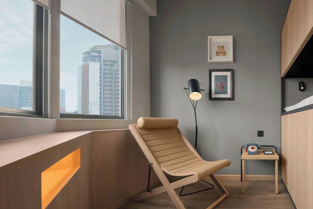A modern room with a large window, a wooden desk, a lounge chair, a floor lamp, and a small table with a telephone.
