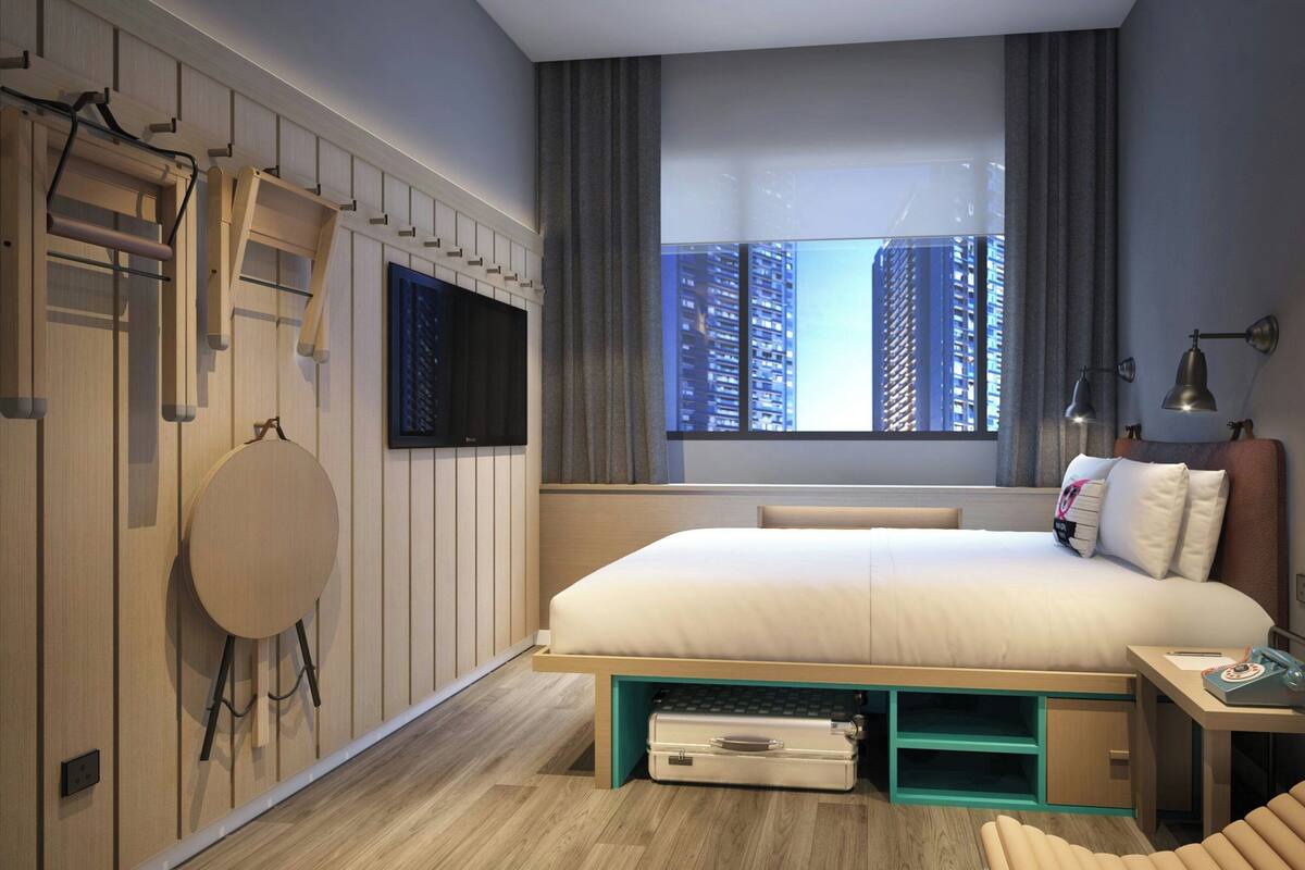 A modern hotel room with a large bed, a flat-screen TV mounted on the wall, a window with a city view, and a desk with a lamp.