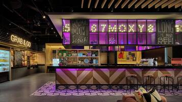 A modern bar area with a neon sign reading "MAYA" and "LONG TIME NO SEE!" The bar has a check-in/out counter and a seating area with stools.