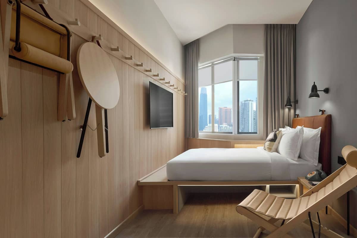 A hotel room with a bed, a TV, a window with a city view, and a wooden chair.