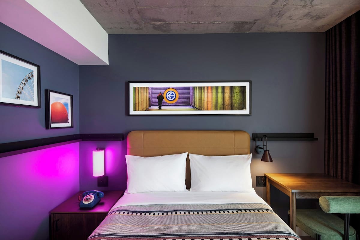 Quarto de hotel com cama, mesa de cabeceira, abajur, quadro emoldurado e obra de arte na parede.