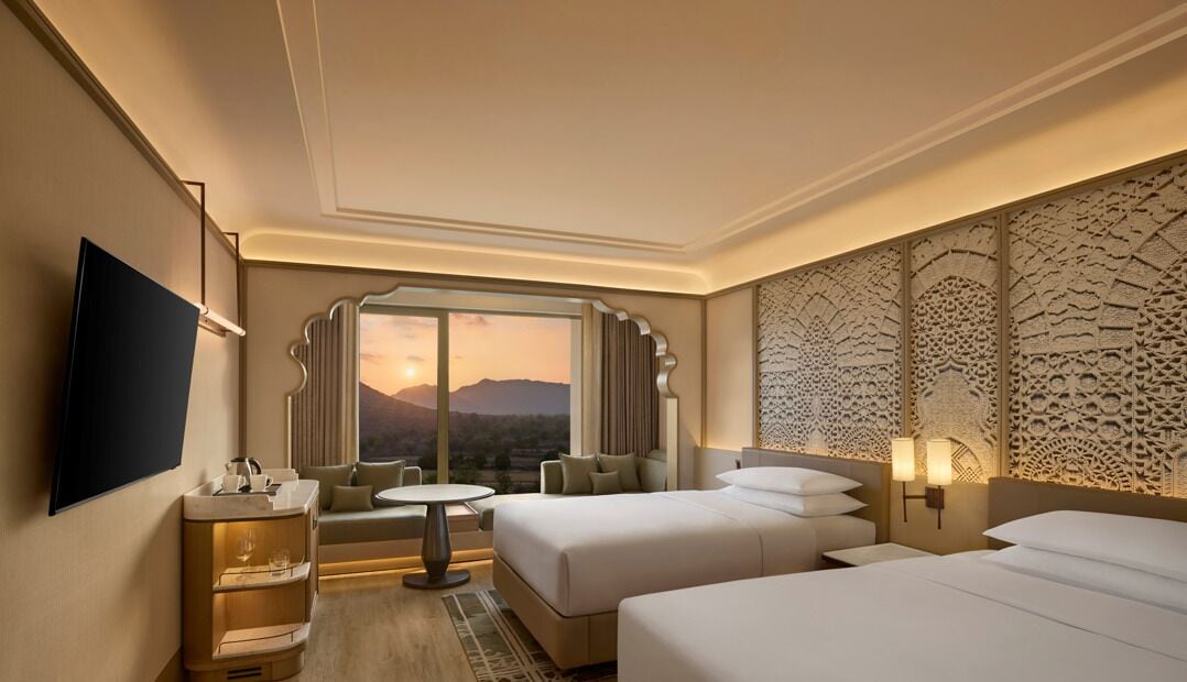 A hotel room with a large window, a flat-screen TV, a sofa, a small table, and two beds.