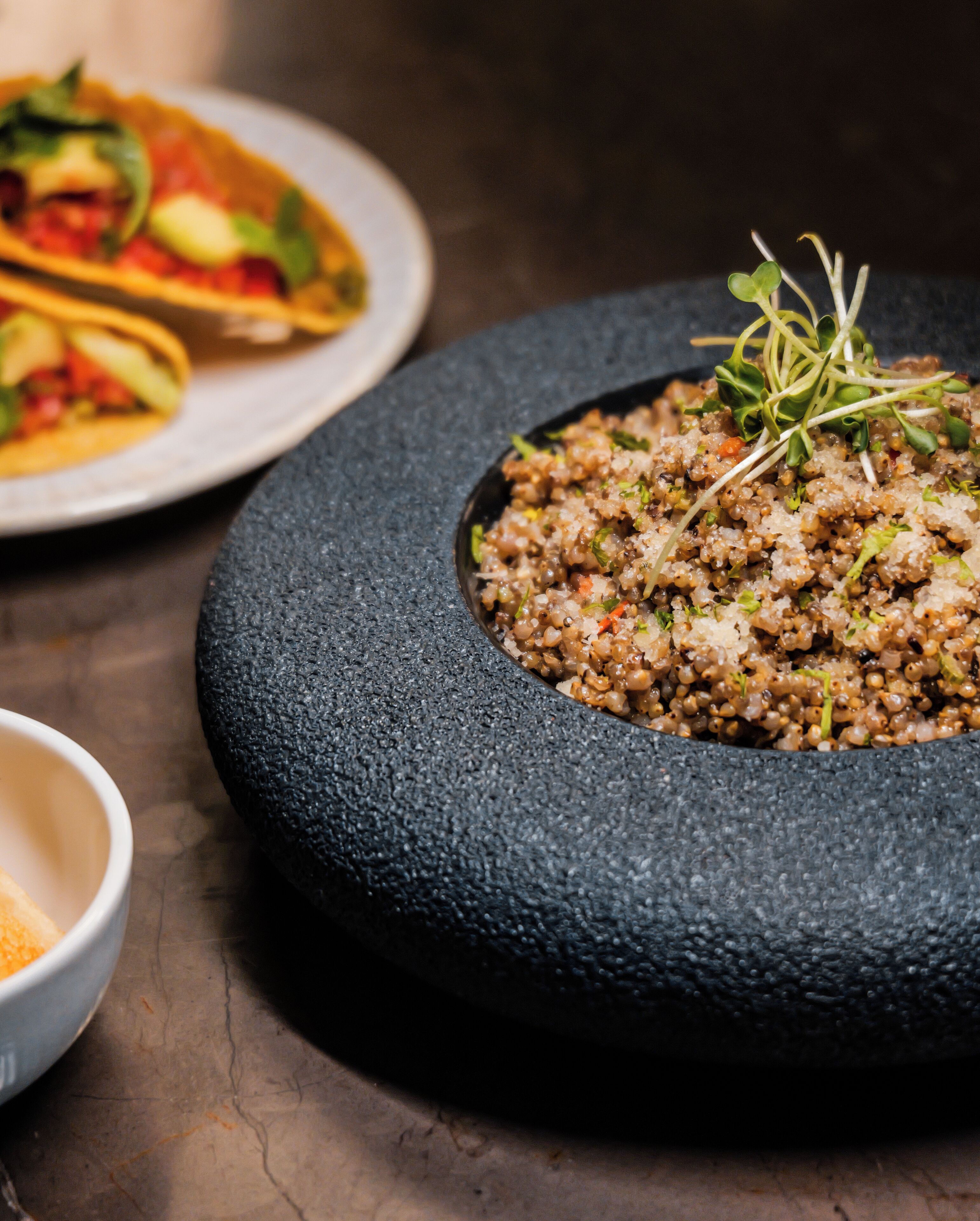 A stone bowl filled with quinoa salad, a plate of tacos, and a small bowl of spices on a dark surface.