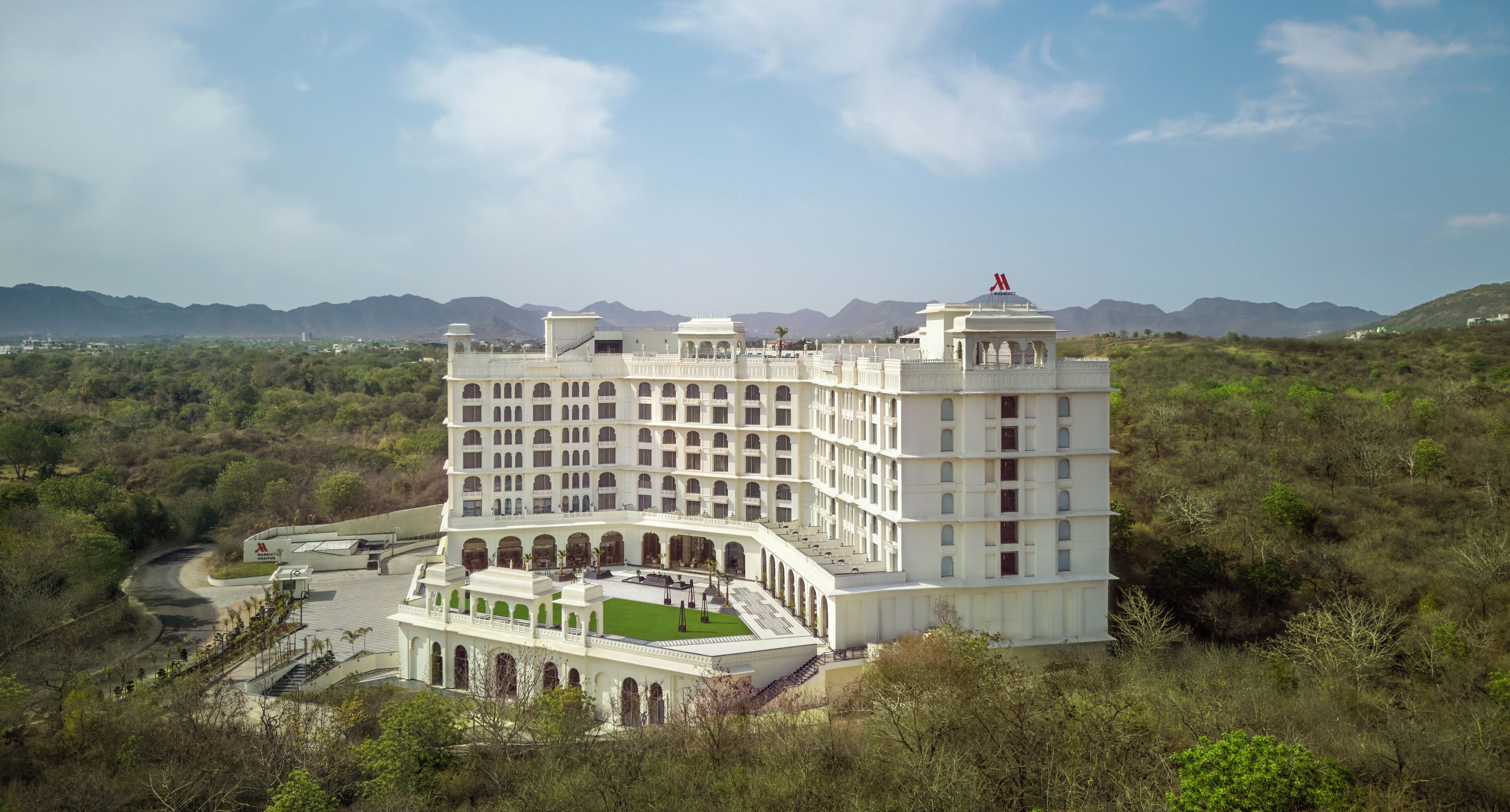 A large, multi-story building with a central courtyard, surrounded by greenery and mountains in the background.