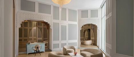 A spacious room with a wooden floor, a blue dresser, and a large mirror. There are three beige armchairs and a small white table with a vase.