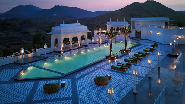 Uma área de piscina com vista para montanhas, um edifício branco com arcos e uma palmeira.